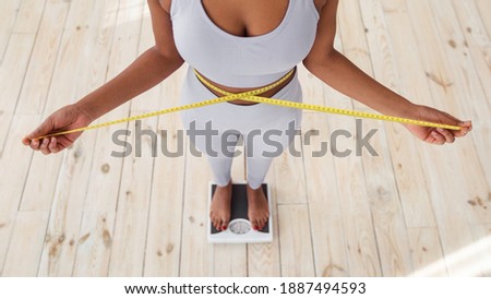 Above view of African American lady measuring her waist with tape, standing on scales indoors, closeup. Young black woman showing results of slimming diet or liposuction, promoting healthy living