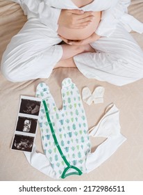 Above View Of Anonymous Woman In White Cozy Pajama, Sitting On Bed With Ultrasound Image Of Womb, Cute Bodysuit And Socks While Caressing Pregnant Belly And Expecting For Baby And Part Of Family