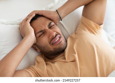 Above View Of Arabic Man Having Headache Suffering From Pain Touching Head Lying In Bed At Home. Sick Guy Having Migraine And Hypertension Symptom. Health Problem Concept