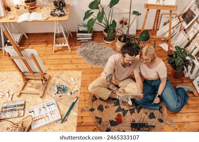 From above view of an artistic creative painter couple sitting on a floor at creative art studio surrounded by paintings and smiling at their artwork. Affectionate artists looking at artwork at studio - Powered by Shutterstock