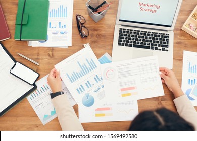 Above View Background Of Unrecognizable Businesswoman Holding Data Charts While Working At Wooden Desk With Documents Laid Out And Words Marketing Strategy On Laptop Screen, Copy Space