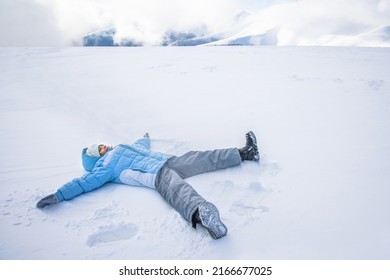 Above View Of Cute Little Girl Making Snow Angel In Fresh Deep Snow. Female Child Wearing Ski Clothes And Skarf Lying, Waving Arms And Legs, Making Angel On Snow In Park. Concept Of Winter Activities.