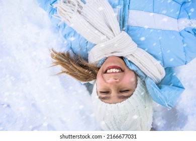 Above View Of Cute Little Girl Making Snow Angel In Fresh Deep Snow. Female Child Wearing Ski Clothes And Skarf Lying, Waving Arms And Legs, Making Angel On Snow In Park. Concept Of Winter Activities.