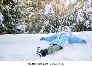 Above View Of Cute Little Girl Making Snow Angel In Fresh Deep Snow. Female Child Wearing Ski Clothes And Skarf Lying, Waving Arms And Legs, Making Angel On Snow In Park. Concept Of Winter Activities.