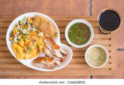 Above View Of Delicous Hornado With Mashed Potatoes With Salad, Tostado And Mote Inside Of A White Plate With Two Smalls Bowls With Green And Pink Sauce And Glass Of Coke