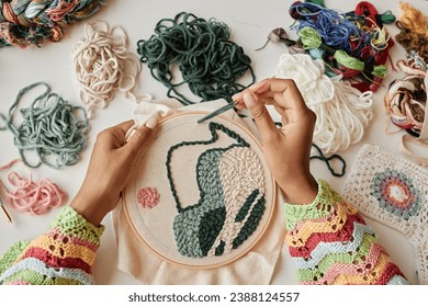Above view of hands of young female designer making creative embroidery on piece of white canvas clamped in hoop