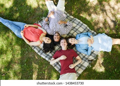 Above View Portrait Of Group Of Friends Lying On Blanket While Enjoying Picnic On Green Grass And Looking Up At Camera, Copy Space