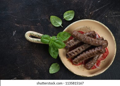 Above view of a serving pan with grilled balkan cevapi sausages with green basil, tomatoes and onion, horizontal shot on a dark brown stone background