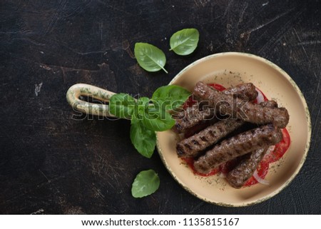 Above view of a serving pan with grilled balkan cevapi sausages with green basil, tomatoes and onion, horizontal shot on a dark brown stone background