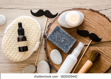 Above View Set Of Various Man Spa Products Things. Natural Wood Disc Background With Flat Sea Stones. Gray Clay Soap Bar, Beard Oil, Moisturizing Cream, Candle Burning. Funny Mustache Shape Figurines.