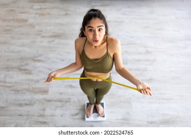 Above view of shocked Indian lady standing on scales, checking her waist measurements, surprised with unexpected results of weight loss program, indoors. Slimming diet and workout concept