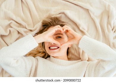 Above view smiling rested lovely blond female looking at camera through shape heart fingers feels refreshed starting new day, good morning, time to get up concept.