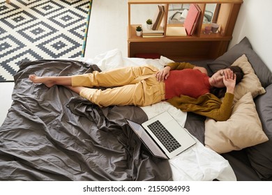 Above View Of Tired Young Woman In Cardigan Lying With Closed Eyes On Bed With Laptop And Holding Hand On Forehead, Work Burnout Concept