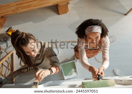 Above view of two young intercultural women with paintroller and paintbrush painting wall of cafe in green during renovation