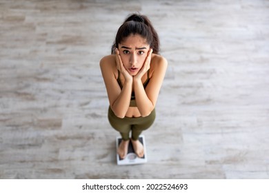 Above view of upset young Indian woman standing on scales, feeling disappointed about her weight loss result indoors. Unsuccessful slimming and dieting concept