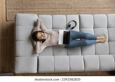 Above view woman rest on sofa with laptop and headphones, put hands behind head lying on couch fall asleep feels carefree looks calm enjoy weekend leisure, fresh conditioned air inside. Relax concept