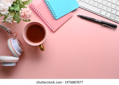Above view of woman workspace with headphone, coffee cup, notebook, keyboard and flowers on pink pastel background.