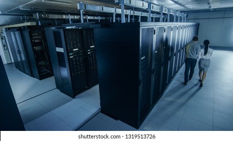 Above Wide Shot Of An IT Admin With A Laptop Computer And Young Technician Colleague Walking Next To Server Racks In Data Center. Running Diagnostics Or Maintenance.