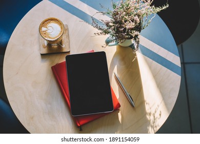 From Above Of Wooden Table Decorated With Bouquet Of Flowers Near Tablet Placed On Red Planner With Pen And Cup Of Aromatic Cappuccino