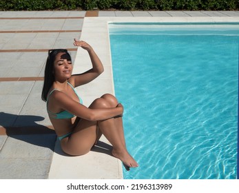 From Above Young Hispanic Woman In Bikini Embracing Knees And Protecting Face From Sun While Sitting On Border Of Pool During Summer Vacation