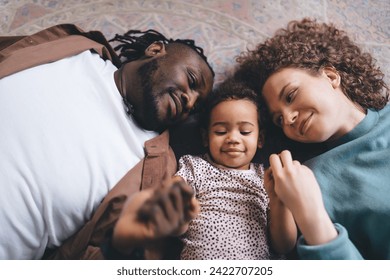 From above of young multiethnic parent in casual clothes with curly hair daughter lying on back over floor holding hands and looking at each other while resting and bonding together in living room - Powered by Shutterstock