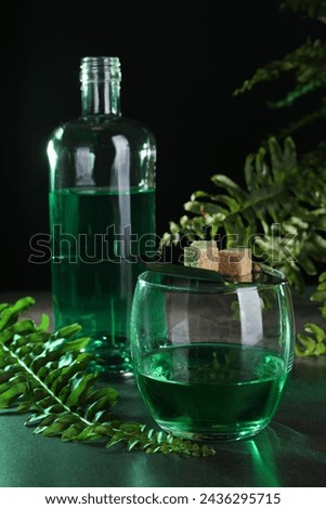 Absinthe in glass, spoon with brown sugar cubes and fern leaves on gray table against black background. Alcoholic drink