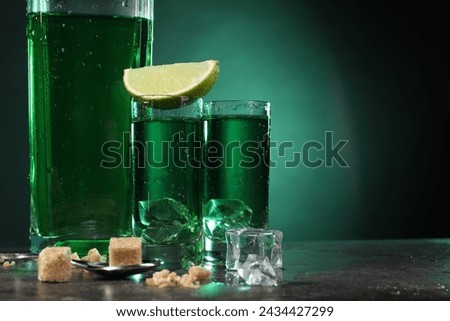 Absinthe in shot glasses with ice cubes, lime, brown sugar and spoon on gray table against green background, space for text. Alcoholic drink