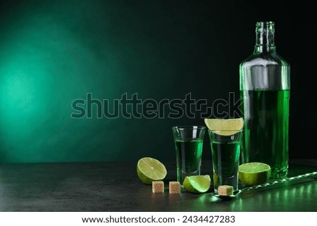 Absinthe in shot glasses, spoon, brown sugar cubes and lime on gray textured table against green background, space for text. Alcoholic drink