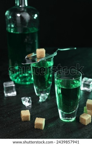 Absinthe in shot glasses, spoon, brown sugar and ice cubes on gray table against dark background, closeup. Alcoholic drink