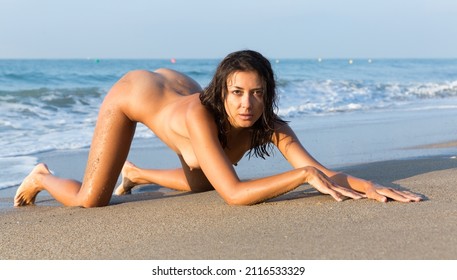 Absolutely naked young woman posing on knees on beach against azure sea
