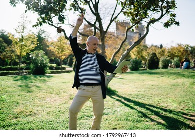 Absorbed 50 Years Old Male Dancing And Looking At Screen Of Tablet While Listening To Music In Earphones On Lawn In City Park