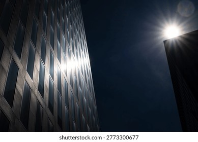 Abstract architectural composition showing two corporate buildings with a brilliant sunburst peeking over the roofline, reflecting light onto a dark window grid pattern in a city business district.