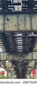 Abstract architectural perspective of the underside of an elevated steel railway bridge creating a striking optical illusion through converging lines, repeating geometric patterns, and dramatic vanishing point composition. Viewed from directly below, the weathered metal framework stretches across the frame, producing a powerful sense of depth, motion, and visual infinity. Riveted steel beams, cross-bracing, and structural elements form an intricate industrial pattern against patches of bright blue sky and surrounding city buildings. The perspective exaggerates the bridge's length, making the structure appear almost endless while showcasing the beauty of engineering and geometric symmetry. This high-resolution image is ideal for architecture, structural engineering, transportation infrastructure, construction, urban design, industrial heritage, abstract backgrounds, geometric compositions, modern city concepts, editorial publications, magazines, presentations, websites, advertising, posters, wall art, educational materials, and creative design. The combination of leading lines, strong perspective distortion, repeating steel textures, and dramatic composition makes this image especially valuable for designers seeking architectural backgrounds, engineering references, futuristic concepts, and visually compelling city infrastructure photography. Perfect for themes of innovation, strength, perspective, precision, transportation systems, and modern metropolitan architecture