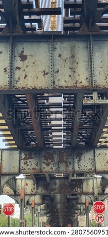 Abstract architectural perspective of the underside of an elevated steel railway bridge creating a striking optical illusion through converging lines, repeating geometric patterns, and dramatic vanishing point composition. Viewed from directly below, the weathered metal framework stretches across the frame, producing a powerful sense of depth, motion, and visual infinity. Riveted steel beams, cross-bracing, and structural elements form an intricate industrial pattern against patches of bright blue sky and surrounding city buildings. The perspective exaggerates the bridge's length, making the structure appear almost endless while showcasing the beauty of engineering and geometric symmetry. This high-resolution image is ideal for architecture, structural engineering, transportation infrastructure, construction, urban design, industrial heritage, abstract backgrounds, geometric compositions, modern city concepts, editorial publications, magazines, presentations, websites, advertising, posters, wall art, educational materials, and creative design. The combination of leading lines, strong perspective distortion, repeating steel textures, and dramatic composition makes this image especially valuable for designers seeking architectural backgrounds, engineering references, futuristic concepts, and visually compelling city infrastructure photography. Perfect for themes of innovation, strength, perspective, precision, transportation systems, and modern metropolitan architecture