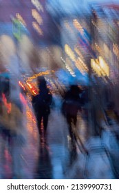 Abstract Background Of Blurred People Figures Under Umbrellas, City Street In Rainy Evening. Light Illumination From Lanterns And Shop Windows. Intentional Motion Blur