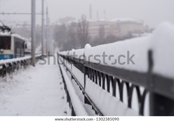 \
Abstract background - snowfall. Intensive falling\
snow on the bridge. A trolley bus and cars are driving along the\
bridge. The handrails and everything around are covered with a\
large layer of snow.