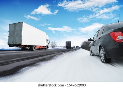 Abstract background with two white box trucks and a black family car on the countryside winter road in motion with snow against a blue sky with clouds