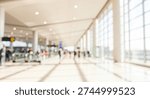 Abstract blurred background of a spacious and bright airport terminal interior. Large windows let in natural light, with silhouettes of people walking in the distance.