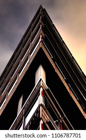 Abstract building pictured in the shape of a triangle. The sky goes from a dark shade on the left to a warmer orange color to the right side of the building. 