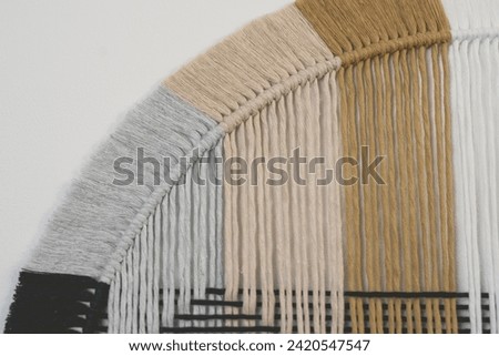 Abstract circular wall decor in white, ochre, beige, gray, and black threads on a clean white wall. Elegant fringe detailing complements the symmetrical design, creating a modern focal point.