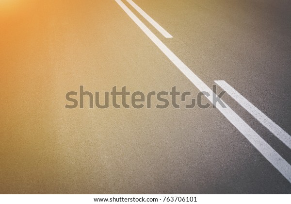Abstract close up image of paved road with\
black tarred surface and white painted dividing lines to indicate\
driving lanes with golden glow from the\
sun
