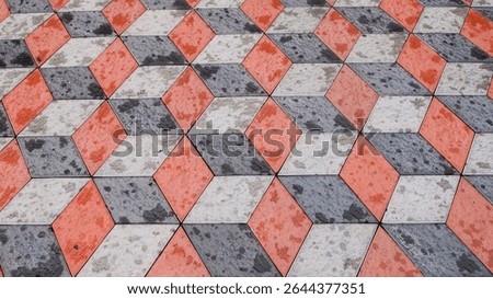 An abstract close-up of a tiled surface featuring a 3D optical illusion pattern composed of red, white, and dark grey rhombus-shaped pavers, some showing water marks or dirt.