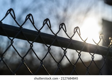 Abstract close-up of the top of a chain link fence with the sun shining through against a blue sky background with blurred silhouettes of trees