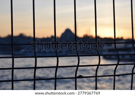 An abstract composition frames the blurred dome and minaret silhouette of the Süleymaniye Mosque at sunset through an out-of-focus metal grid fence. The warm sunset tones bleed softly through the bars in an impressionistic take on the Istanbul skyline.