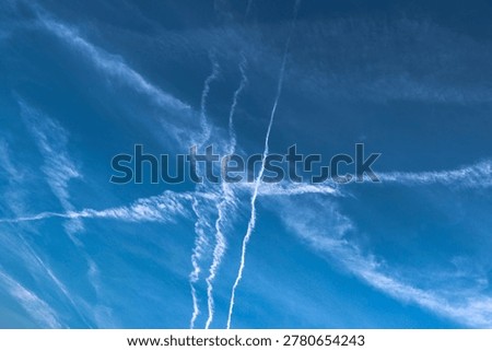 Abstract composition of intersecting airplane contrails and soft clouds across a vivid blue sky.