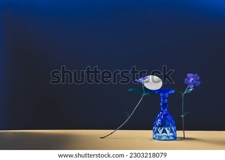 Abstract composition with periwinkle flowers, upside down blue wine glass and white stone.
