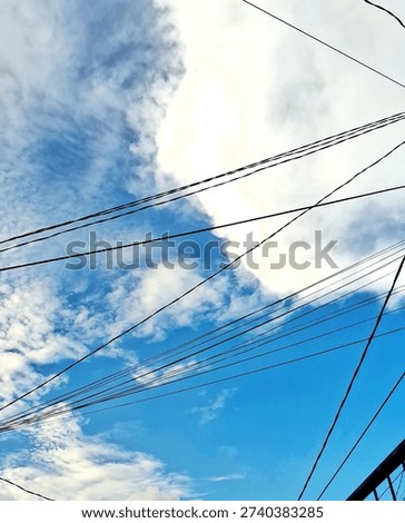 Abstract composition of tangled black electrical wires crisscrossing against a bright blue sky and large white fluffy clouds.