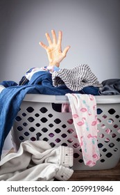 Abstract Concept Image With A Woman's Hand Coming Out Of A Basket Full Of Clothes For Laundry. She Is Overwhelmed And Bored With Housework And Needs Help. Repetitive Never Ending Work