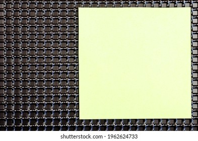 An abstract geometric composition of black repeating squares and a sheet of paper. A light green blank piece of paper on top of a pattern of identical shapes. Top view. Copy space.