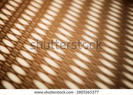 Abstract image featuring light and shadow patterns formed by a perforated surface, creating elongated oval shapes on a textured background